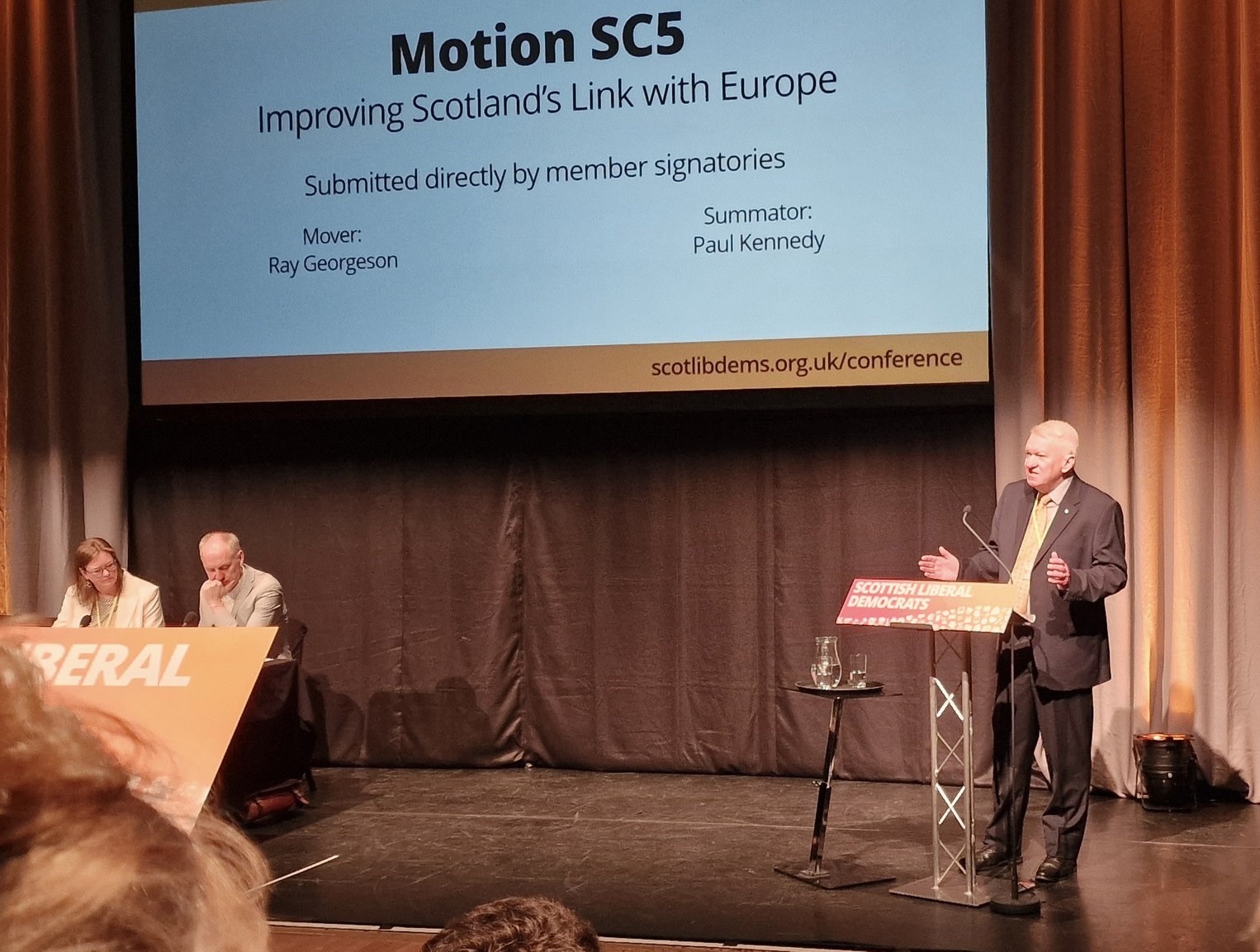 A photo of Ray standing and talking on a podium with the sign Scottish Liberal Democrats