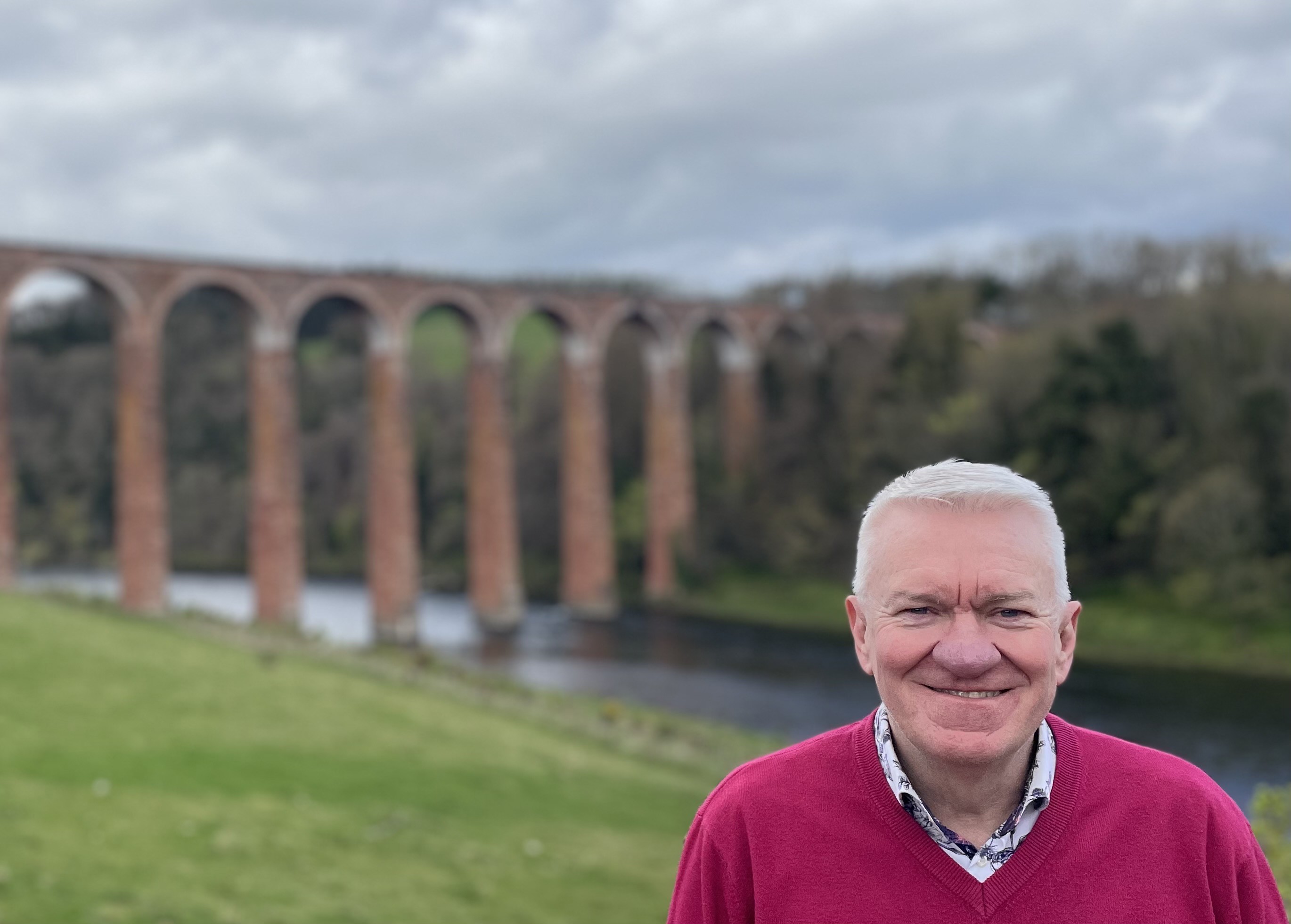 A photo of Ray smiling in the foreground, with an aqueduct in the background