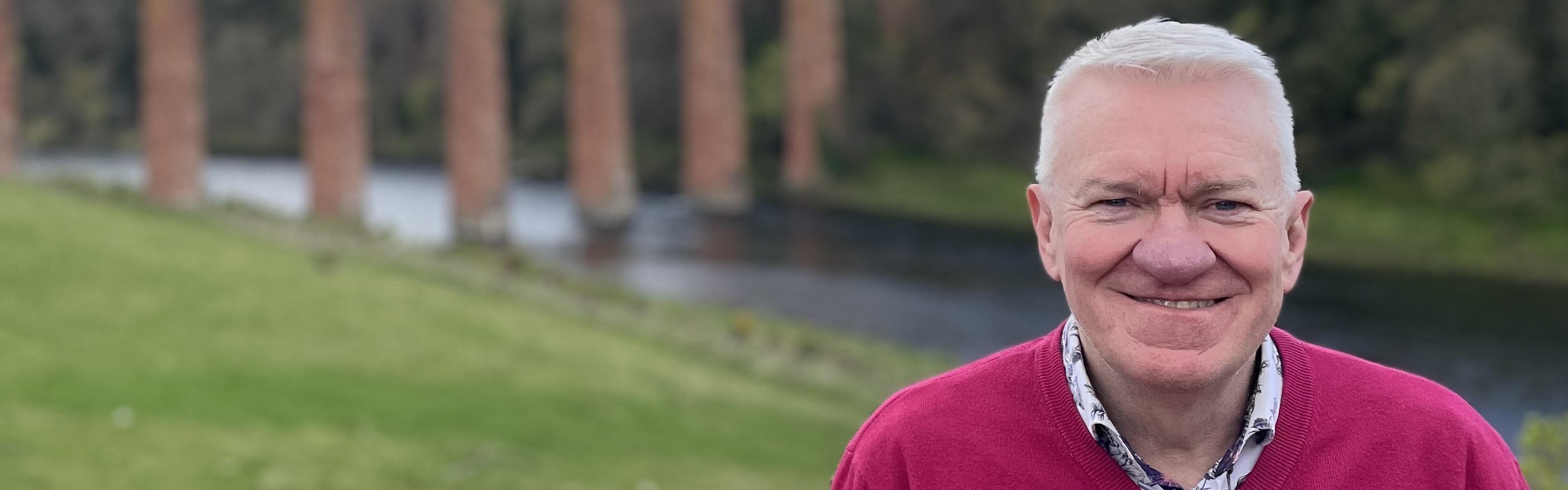 A photo of Ray smiling in the foreground, with an aqueduct in the background
